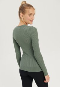 Grüner gerippter Pullover mit langen Ärmeln, rundem Halsausschnitt und taillierter Silhouette, kombiniert mit schwarzen gerippten Leggings.