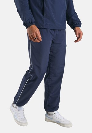 Man draagt marineblauwe trainingsbroek met witte zijstreep, witte sokken en witte sneakers, staand tegen een effen achtergrond.