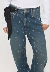 Blue denim jeans featuring silver eyelet embellishments, a relaxed fit, and a black fabric tie at the waist.