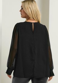 Schwarze Bluse mit durchsichtigen Puffärmeln, strukturellem Smocking an den Bündchen, Schlüsselaugen-Details im Rücken und lockerer Passform.