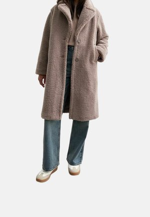 Femme portant un manteau teddy beige, un jean bleu à jambes larges et des baskets blanches, debout avec une main dans la poche du manteau sur un fond blanc.