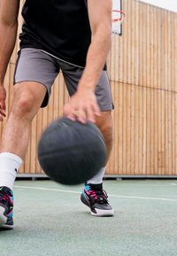 Juodas krepšinis su tekstūruota paviršiumi, laikomas asmens su pilkais šortais ir juoda marškinėliais, avinčiu spalvingus sportbačius, krepšinio aikštelėje su mediniu fono.