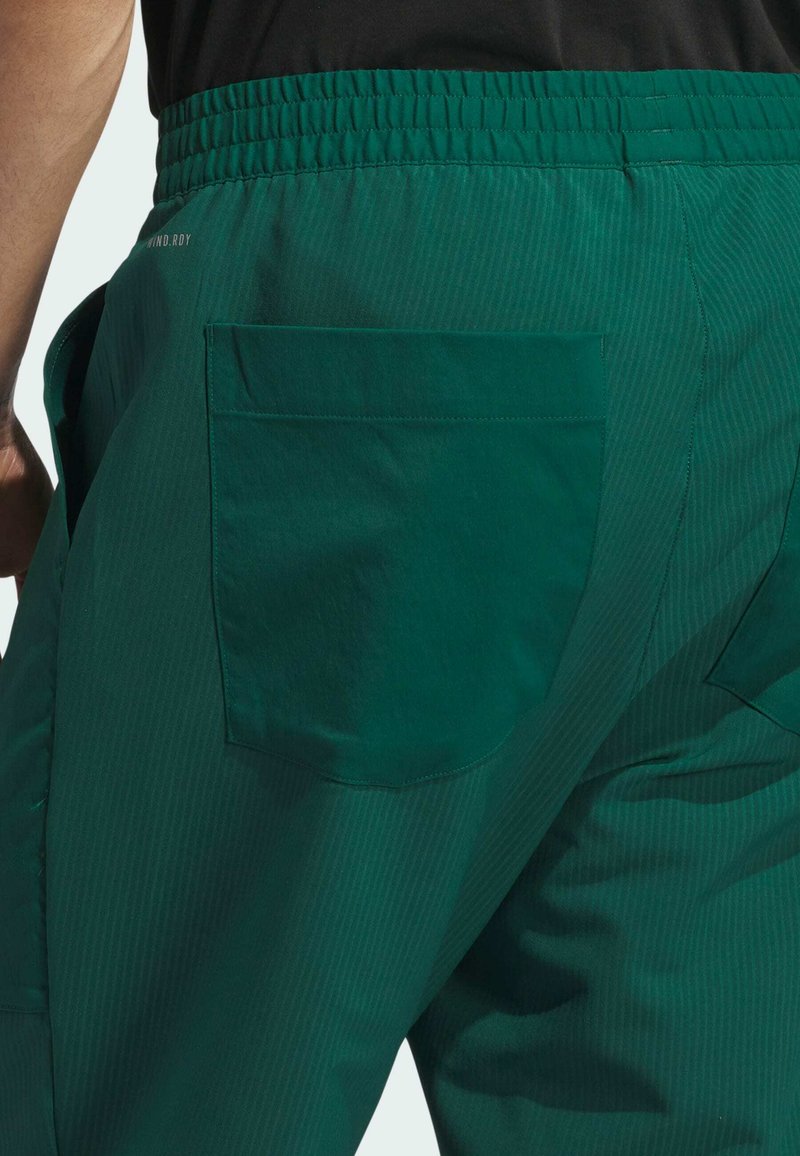 Grüne Hose mit strukturierten Oberflächen, elastischem Bund und einer Gesäßtasche. Der Stoff hat vertikale Streifen und eine glatte Oberfläche.
