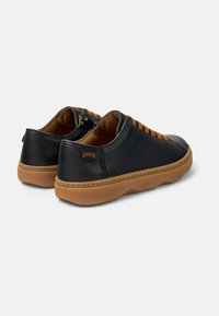 Zapatillas de cuero negro con cordones marrones y suelas de goma. Presentan una superficie texturizada y un pequeño logo en el talón. Forma redondeada.