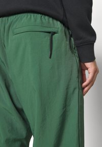 Zelené, lehké kalhoty se hladkým povrchem, s kapsou na zip na zadní straně a černými šňůrkami. Viditelný elastický pas.