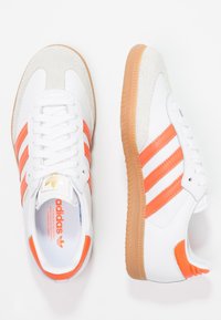 Vita sneakers med orange ränder, mockatåhättor och texturerade sulor i gummimaterial. Har snören och logotyp på tungan.