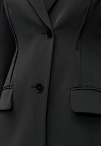 Gros plan d'un blazer noir sur mesure avec deux boutons noirs et deux poches à rabat à l'avant.