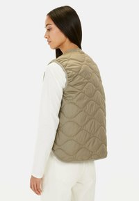 Beige Steppweste mit hohem Kragen und ärmellosem Design, ausgestattet mit einem wellenförmigen Muster und grauen Besätzen. Tragen Sie sie über einem weißen Langarmshirt.