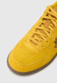 Žlutá semišová teniska se zaoblenou špičkou, perforovanými detaily a hnědými akcenty. Má ploché tkaničky a texturovanou gumovou podrážku.