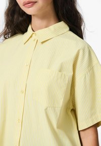 Geel gestreept shirt met een kraaglijn, korte mouwen en een borstzak aan de linkerzijde. De stof lijkt glad te zijn, met dunne witte strepen.