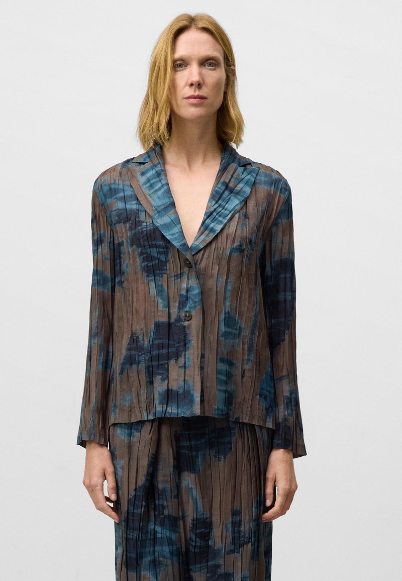 Vrouw met schouderlang blond haar, gekleed in een geplooide blauwe en bruine gestreepte lange mouwen jas en een bijpassende rok tegen een effen achtergrond.