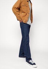 Hombre vestido con jeans azules, zapatillas navy con cordones blancos, camiseta gris y chaqueta marrón de botones, de pie con las manos en los bolsillos.
