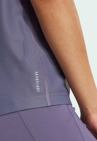 Lila Sportoberteil mit strukturiertem Material und "AEROREADY"-Logo, kombiniert mit glatten lilafarbenen Hosen, die Nähte und Konturen betonen.