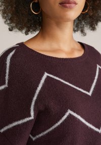 Bordeauxfarbener Pullover aus weichem, strukturiertem Stoff mit einem weißen Zickzackmuster und einem weiten, runden Ausschnitt.