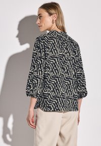 Blusa nera e crema a fantasia con maniche a tre quarti, scollatura arricciata e leggero svaso all'orlo, indossata sopra pantaloni chiari.