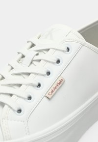 Weiße Calvin Klein Sneaker mit weißen Schnürsenkeln, Metallösen und sichtbarem Markenetikett an der Seite in der Nähe der Schnürsenkel.