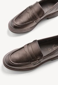 Mocasines de cuero marrón con un acabado texturizado, que presentan una costura frontal elevada, tira tipo penny y un tacón bajo en madera. El interior es perforado.