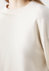 Jasbeżowy sweter z prążkowanym dekoltem i gładką fakturą, o luźnym kroju i nieco opadających szwach ramion.