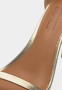 Κοντινό πλάνο ενός χρυσού πέδιλου με ψηλό τακούνι, λεπτό λουράκι και ταμπά πάτο με την ετικέτα "Call It Spring Vegan".