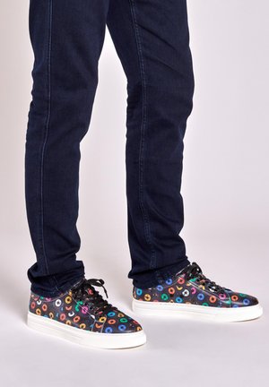 Multicolored sneakers with circular patterns, black synthetic upper, white rubber sole, and black laces, paired with dark denim jeans.
