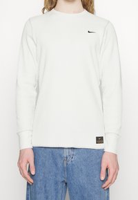 Vit långärmad ribbad tröja med rund hals, med en svart Nike-logotyp på bröstet och en märkeslapp vid fållen.