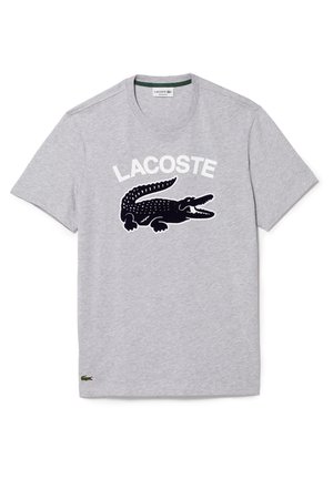 T-shirt gris à manches courtes avec un grand crocodile noir et le texte blanc "LACOSTE" sur le devant, ainsi qu'un petit logo crocodile vert près de l'ourlet.