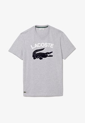 T-shirt gris à manches courtes avec un grand crocodile noir et le texte blanc "LACOSTE" sur le devant, ainsi qu'un petit logo crocodile vert près de l'ourlet.