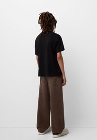 Black cotton t-shirt with a relaxed fit, short sleeves, paired with brown wide-leg pants and beige sneakers, viewed from the back.