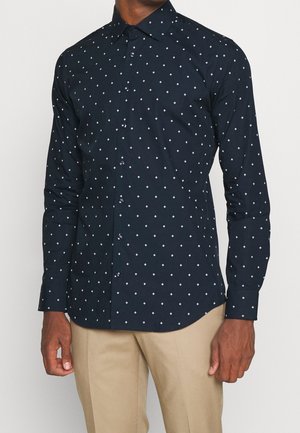 Mann trägt ein dunkelblaues Hemd mit kleinen weißen Rautenmuster und beigen Hosen, steht vor einem einfarbigen Hintergrund.