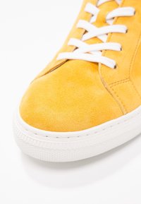 Baskets en daim jaune avec lacets blancs et semelle en caoutchouc blanche épaisse, vues en gros plan sur les détails avant et latéraux.