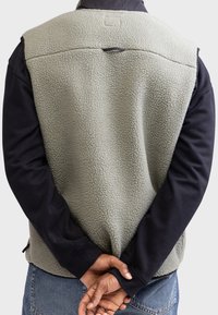 Light green fleece vest with a textured surface, contrasting black sleeves, and a back hook detail. Worn over a dark long-sleeve shirt.