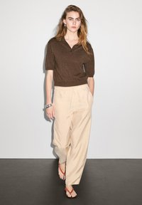 Hnědý polokošile s krátkým rukávem a strukturovaným povrchem, doplněná volnými béžovými kalhotami. Model má na sobě červené žabky, stříbrný náramek a dlouhé vlasy.