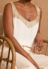 Krémový top na ramínka s výstřihem do V zdobený zlatými flitry. Hladká tkanina, volný střih, v kombinaci se světle béžovými kalhotami.