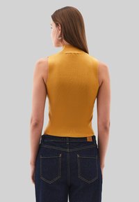 Mosterdkleurige mouwloze top met hoge kraag, textuurstof, aansluitend ontwerp, gecombineerd met donkere spijkerbroek met gele stiksels.