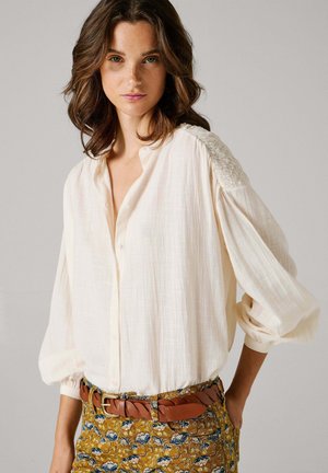 Kremfarvet bluse med knaplukning, strukturerede skuldre og let puffede ærmer. Kombineret med mønstrede sennepsfarvede bukser og et brunt flettet bælte.