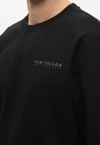 Fekete pulóver kerek nyakkal, pamutból készült. Az elején, a bal mellkason egy szürke "TOM TAILOR" logó található. Sima textúrájú, laza stílusú.