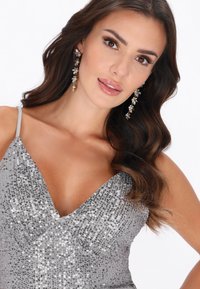 Silbernes Pailletten-Kleid mit tiefem V-Ausschnitt und dünnen Spaghettiträgern, mit figurbetonter Silhouette und glatter Textur. Lange, dekorative Ohrringe.