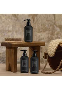 Tre bottiglie nere opache con erogatori a pompa su un tavolo di legno, accompagnate da un pennello in setole naturali e un panno bianco texturizzato.