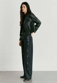 Mujer joven con chaqueta de cuero negra, camiseta negra y vaqueros negros de pierna ancha con un patrón lateral de pequeñas tachuelas plateadas.