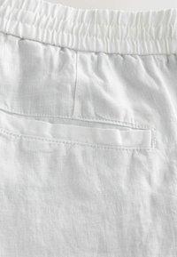 White linen fabric featuring a textured surface, with an elastic waistband and a small back pocket stitched with matching thread.