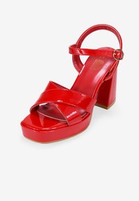 Sandal de plataforma de charol rojo con tacón grueso y tira en el tobillo. Presenta correas cruzadas sobre los dedos y un suave forro interior.