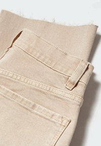 Tela de mezclilla beige con un borde deshilachado, que presenta un lazo de cinturón y costuras en los bolsillos traseros. Textura suave con acentos de costura ligera.