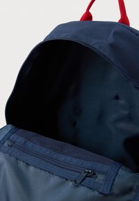 Ναυτικό μπλε σακίδιο με κόκκινες λαβές, με κεντρικό διαμέρισμα με φερμουάρ και λεπτομέρεια δίχτυ στην επάνω άκρη. Ομαλή υφή ορατή.