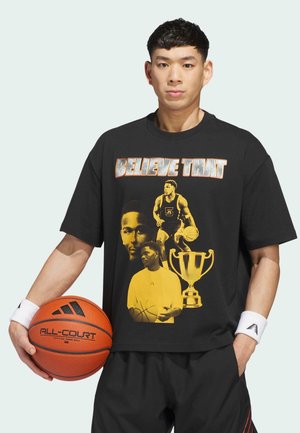 Young man holding basketball wearing black shirt with basketball player images, trophy, and "BELIEVE THAT" text in bold letters.