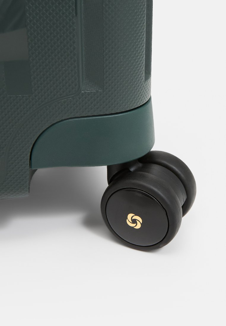 Grüner Hartschalenkoffer mit strukturiertem Oberflächenmuster, schwarzen Gummirollen und goldfarbener Logo-Details an der Rolle, die Haltbarkeit und Mobilität betonen.