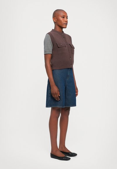 Gilet smanicato marrone a maglia con chiusura a bottoni e due tasche frontali, indossato sopra una camicia a righe bianco e nere, abbinato a pantaloni corti in denim e ballerine nere.