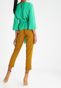 Blusa verde a maniche lunghe con vita legata e scollo a chiave, abbinata a pantaloni giallo senape a sigaretta e stivaletti beige con tacco.