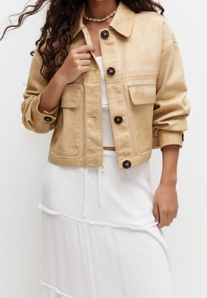 Femme portant une veste courte beige à gros boutons foncés, un haut blanc côtelé, une jupe blanche à volants et un collier en coquillage.