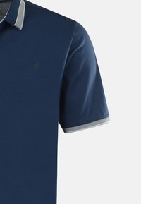 Mörkblå pikétröja med en strukturerad tyg. Har en kontrasterande grå krage och mudd, samt en liten, präglad logotyp på bröstet.