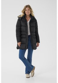 Abrigo acolchado negro con capucha, que cuenta con un cierre de botones, un diseño acolchado, bolsillos laterales y un corte relajado, combinado con jeans acampanados y zapatillas.